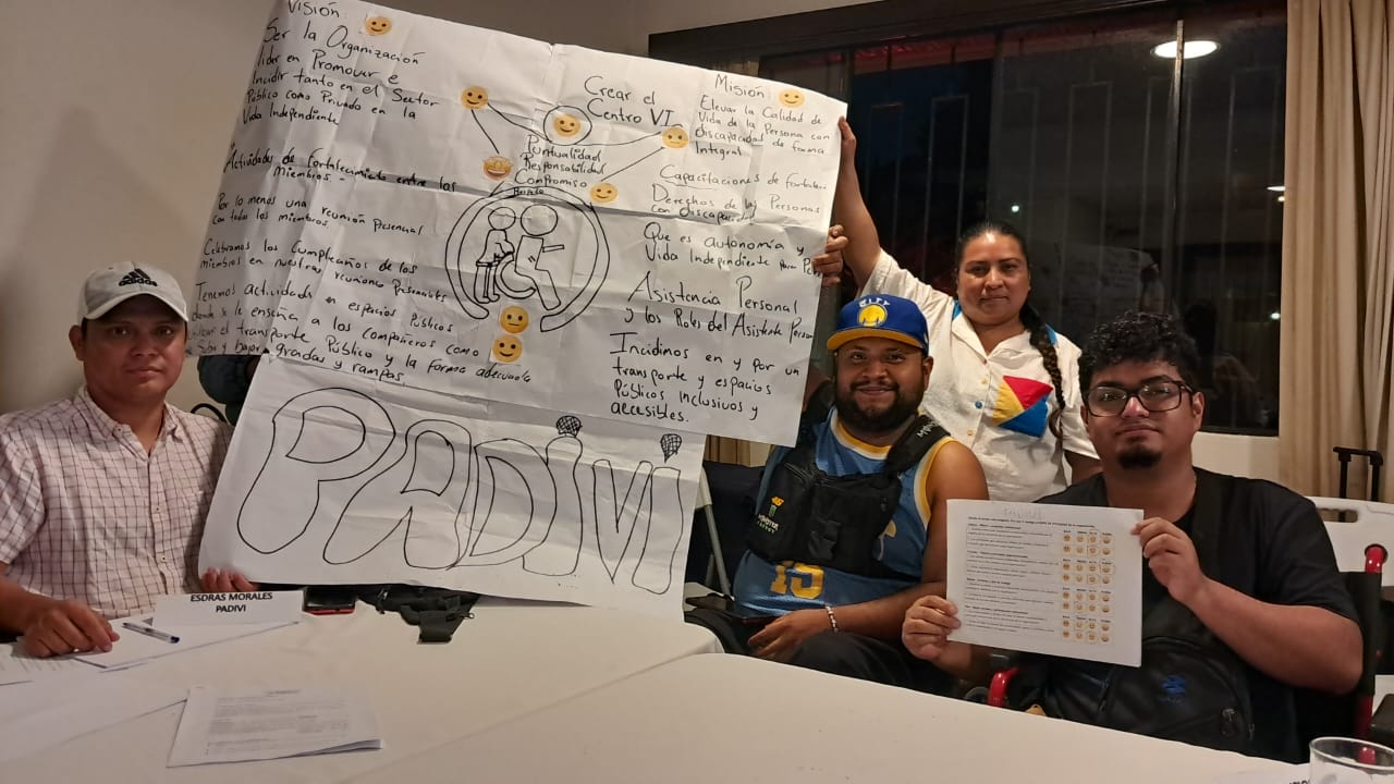 Socialización de los estatutos de las asociaciones Los representantes de las asociaciones compartieron la misión, visión, comisiones y actividades que realizan. Texto alternativo: Cinco personas en sillas de ruedas muestran carteles con dibujos, emojis y textos sobre inclusión. Uno de los carteles tiene el acrónimo “PADIVI”. El ambiente es colaborativo.