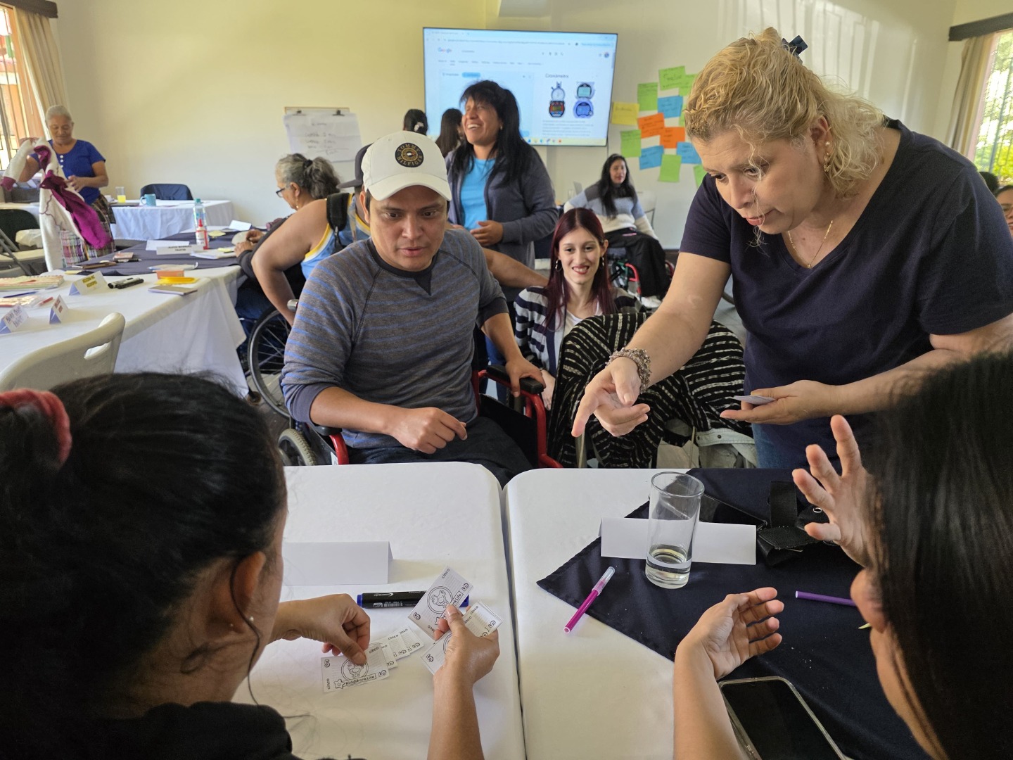 Una de las dinámicas para la promoción de la equidad fue la asignación de roles, como padres, madres, empresas con vacantes laborales, guardería, tienda de verduras, tienda de limpieza, bancos, y venta de leña. Cada padre y madre tenía la tarea de ir buscar un empleo, dejar a sus hijos a la guardería, comprar los alimentos, artículos de limpieza y leña en un tiempo establecido. Esto fomento la empatía y la asignación de roles de forma equitativa, sobre todo para las mujeres que se dedican al hogar y se limitan de una participación en las asociaciones. Texto alternativo: Varias personas, algunas en silla de ruedas, están sentadas alrededor de mesas en una sala bien iluminada. Una mujer señala algo mientras conversa con el grupo, generando un momento de intercambio activo. Sobre la mesa hay materiales impresos, bolígrafos y tarjetas con nombres. Al fondo, se ve una pantalla con dibujos y una pared con notas adhesivas de colores, lo que sugiere que están en una dinámica de aprendizaje colaborativo.