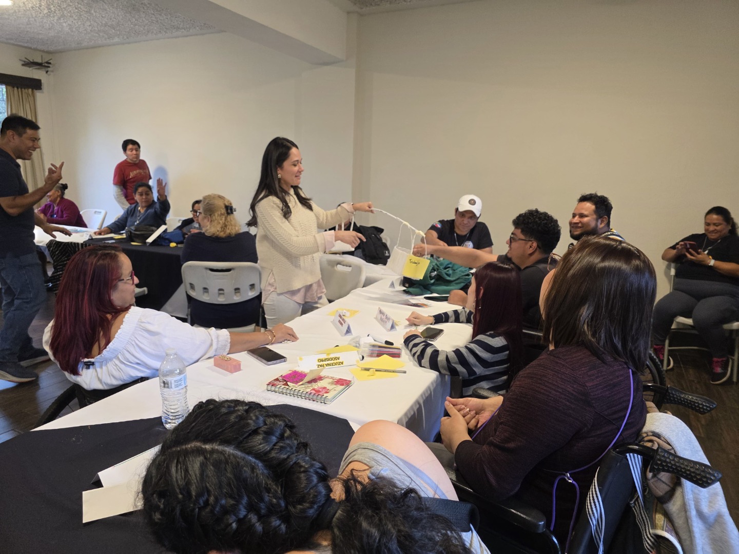 Uno de los puntos durante el taller, fue identificar y reflexionar sobre aquellas acciones que tienen mayor relevancia en el diario vivir. Por ejemplo, el trabajo, dejando en el olvido el cuidado personal. Texto alternativo: Una mujer entrega una bolsa amarilla a una persona sentada, mientras otras observan con atención y alegría. Están en una sala con mesas, cuadernos y tarjetas de nombre. Participan personas en silla de ruedas. El ambiente es cálido, organizado y participativo.