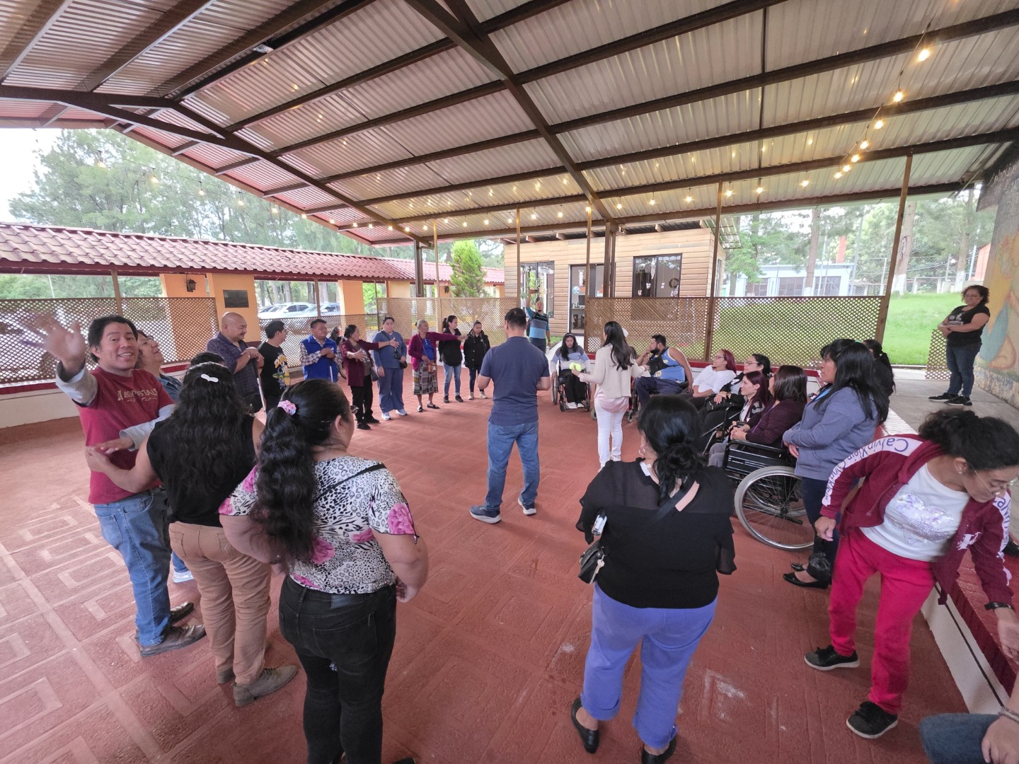 A través de una dinámica grupal, cada participante expresó acciones concretas para promover la equidad en las asociaciones. Texto alternativo: Un grupo diverso de personas se reúne bajo un techo metálico en un área abierta con piso de cerámica roja. Algunas personas están de pie y otras en silla de ruedas. El ambiente es cálido y participativo, decorado con luces colgantes y un mural colorido en la pared. Se percibe una atmósfera de inclusión y convivencia, como si se tratara de una actividad recreativa o social en comunidad.