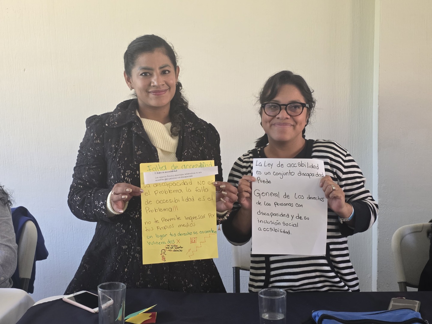Se promovió la participación segura e inclusiva para cada socio. Texto alternativo: Dos personas están sentadas en una mesa, cada una sosteniendo un cartel escrito a mano. El cartel amarillo dice que la falta de accesibilidad es el verdadero problema, no la discapacidad. El cartel blanco habla sobre la ley de accesibilidad y los derechos de las personas con discapacidad. El ambiente es de reflexión y empoderamiento, en un espacio que promueve el diálogo sobre derechos y barreras sociales.